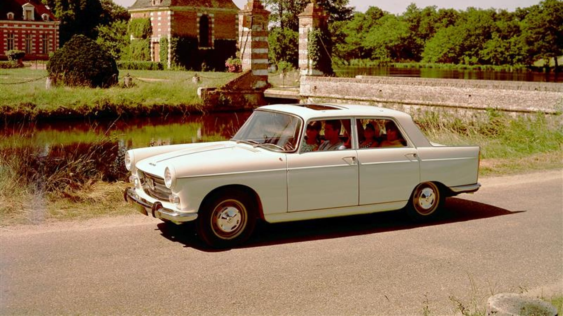 PEUGEOT 404 na wiejskiej drodze przed czerwono ceglanym dworem i w otoczeniu zieleni.